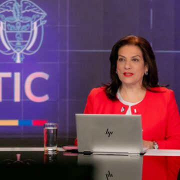 Alexandra Falla Zerrate in a red jacket seated at a studio desk with a laptop, a MinTIC logo on the screen behind her