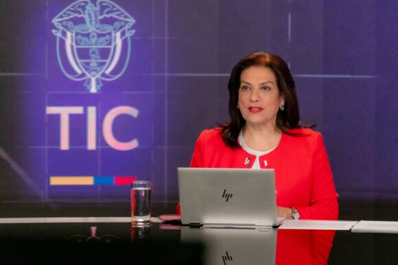 Alexandra Falla Zerrate in a red jacket seated at a studio desk with a laptop, a MinTIC logo on the screen behind her