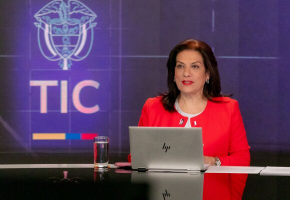 Alexandra Falla Zerrate in a red jacket seated at a studio desk with a laptop, a MinTIC logo on the screen behind her